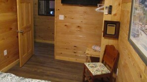 A wooden cabin room with a bed, a small table, a chair, a television, and a framed picture on the wall. - A cozy new cabin in rural setting with a hot tub on porch. Beautiful rural view. (Albany)
