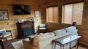 A cozy living room with a fireplace, a sofa, a coffee table, and a TV mounted on the wall. - A cozy new cabin in rural setting with a hot tub on porch. Beautiful rural view. (Albany)