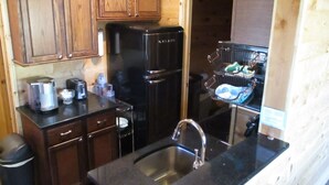 A compact kitchen with wooden cabinets, a black refrigerator, a sink, and a dish rack. - A cozy new cabin in rural setting with a hot tub on porch. Beautiful rural view. (Albany)