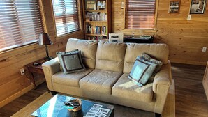 A cozy living room with a beige sofa, a glass-top coffee table with photos, a wooden bookshelf, and wooden walls. - A cozy new cabin in rural setting with a hot tub on porch. Beautiful rural view. (Albany)