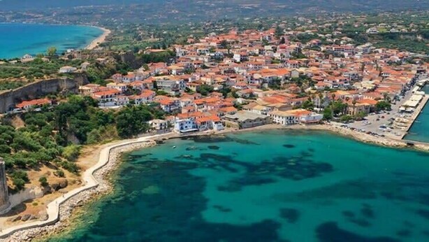 Vista aérea de un pueblo costero con una fortificación destacada, un puerto deportivo y una zona residencial densamente poblada con casas de tejados rojos.