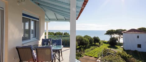 A balcony with a table and chairs, overlooking a coastal landscape with a clear view of the sea and greenery.