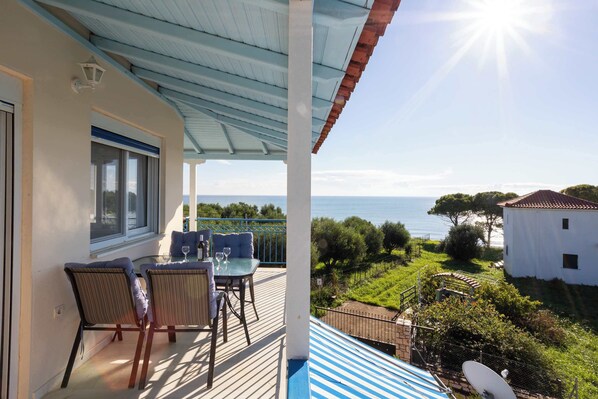 A balcony with a table and chairs, overlooking a coastal landscape with a clear view of the sea and greenery.