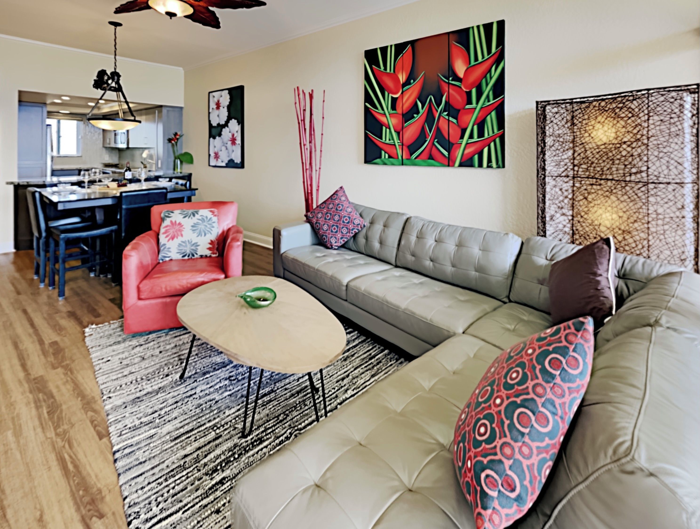 Ein modernes Wohnzimmer mit grauem Sofa, rotem Sessel und Couchtisch. Es gibt einen Essbereich mit Tisch und Stühlen, und eine Küche im Hintergrund.