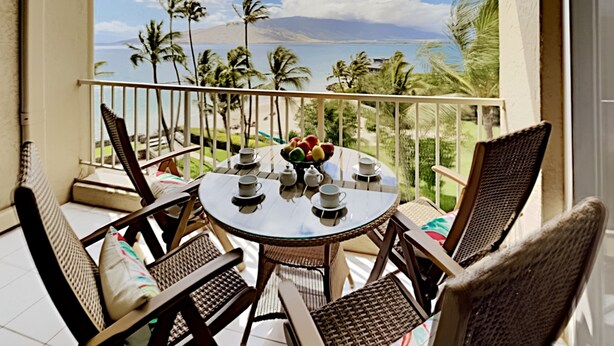 Een balkon met een ronde tafel en rieten stoelen, met uitzicht op een strand met palmbomen en een heldere lucht.
