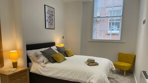 Une chambre à coucher avec un lit, des lampes de chevet, une chaise et une fenêtre donnant sur des immeubles.