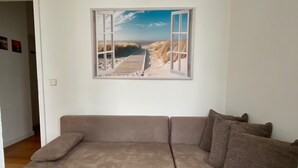 A beige sofa with a cushion, a brown cushion, and a framed picture of a beach scene with dunes and a wooden boardwalk. - Vacation apartment Villa am Meer - Experience Stralsund (Stralsund)