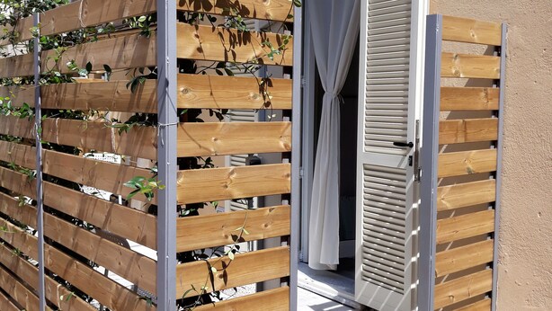 A wooden fence with climbing plants, a sliding glass door, and a tiled patio area. - Flatty Alassio - Three Dots (Alassio)