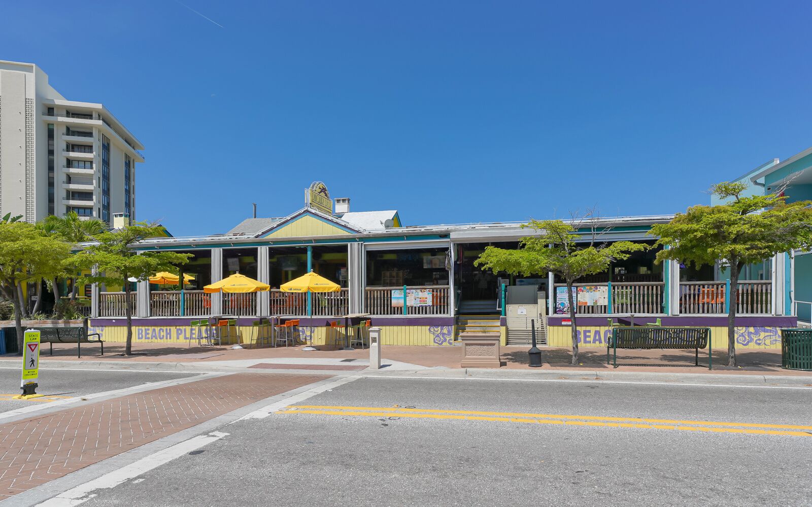 A beachside restaurant with outdoor seating, a clear sky, and a pedestrian crossing.