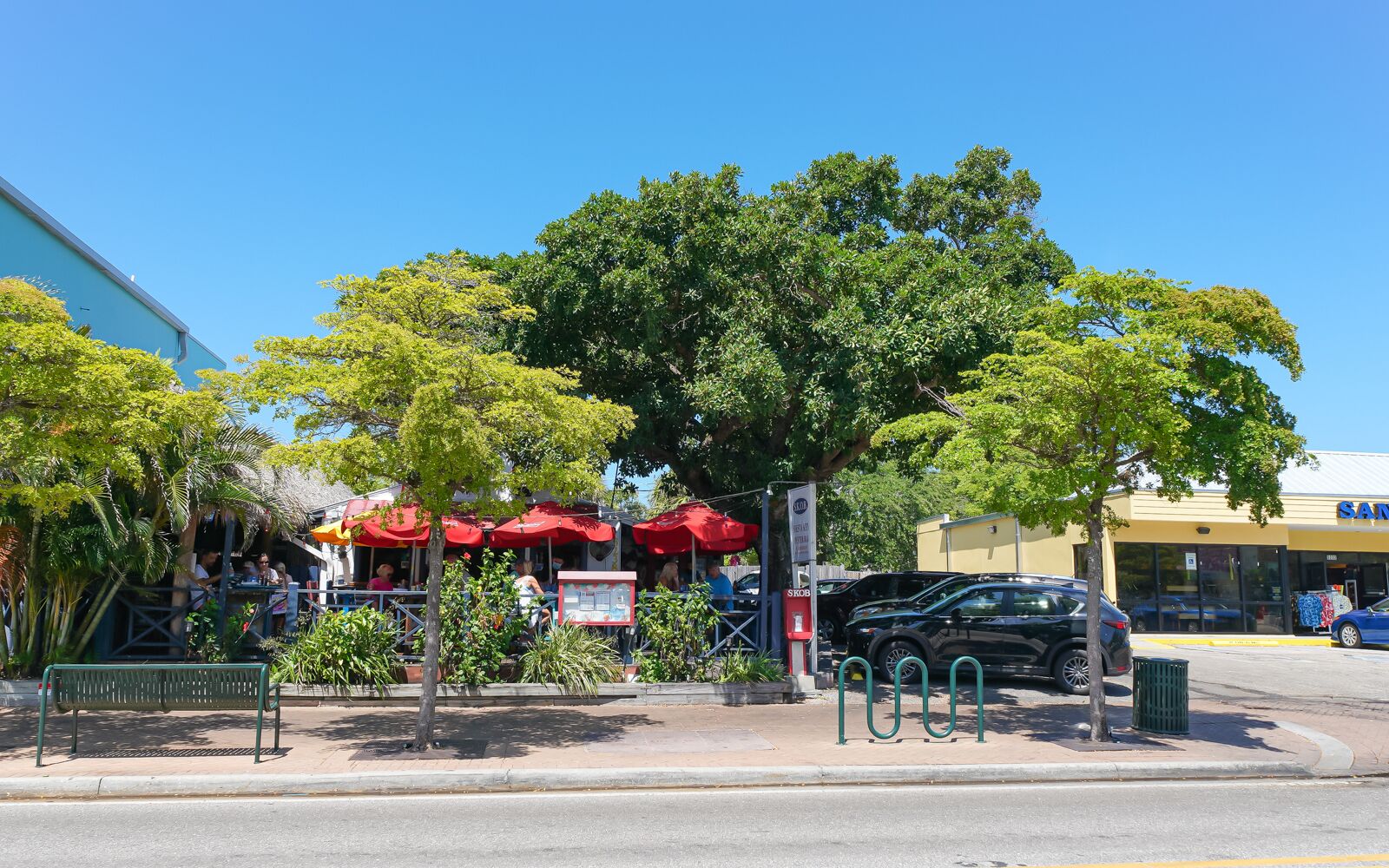A street with outdoor seating, a parking area, and a building with a 'S' sign.