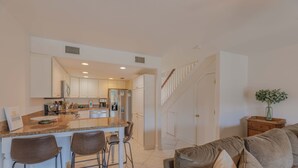 A modern kitchen with white cabinets, a central island with bar stools, and a staircase leading to another level. - Cozy Coastal Beach Villa (Pensacola Beach)