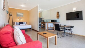A compact living space with a bed, a red sofa, a small wooden table, a TV, a kitchen area, and a dining table with chairs.
