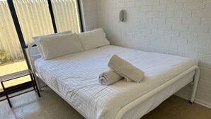 A neatly made bed with white bedding and a rolled towel, a small wooden nightstand, and a white brick wall. - Windsurfer Beach Chalet 3 - No frills holiday vibe (Lancelin)