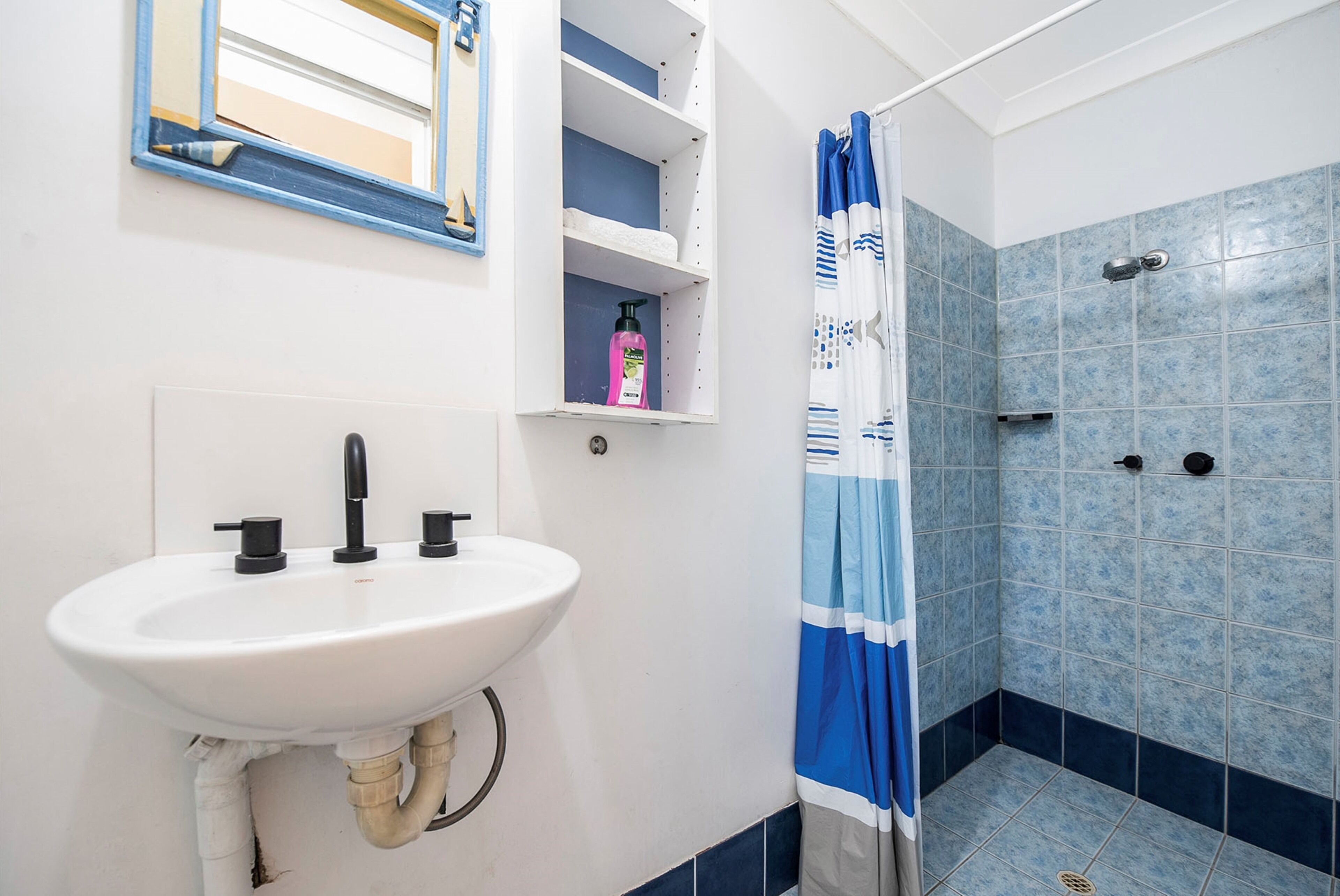 Een badkamer met een witte wastafel, een spiegel, een plank met toiletartikelen en een douche met blauwe tegels.