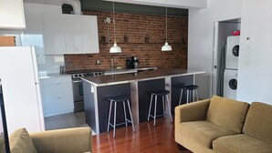 A compact kitchen with a brick wall, white cabinets, a central island with stools, and a washing machine in the background. - Huge terrace - Downtown condo - Free parking - Near Old Port (Montréal)