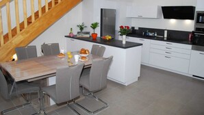 A modern kitchen with white cabinets, a wooden staircase, and a dining area with a table and chairs. - Appartement T3 Avec Terrasse et Accès Direct à la Plage (Fréhel)