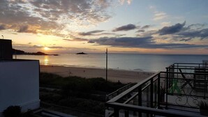 A beach view from a balcony with a railing, a clear sky, and the sun setting over the water. - Appartement T3 Avec Terrasse et Accès Direct à la Plage (Fréhel)