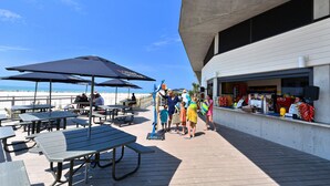 An outdoor seating area with tables and umbrellas, a beach in the background, and a building with a white facade. - Siesta Cooler: Only Minutes Walk to Siesta Beach! (Siesta Key)