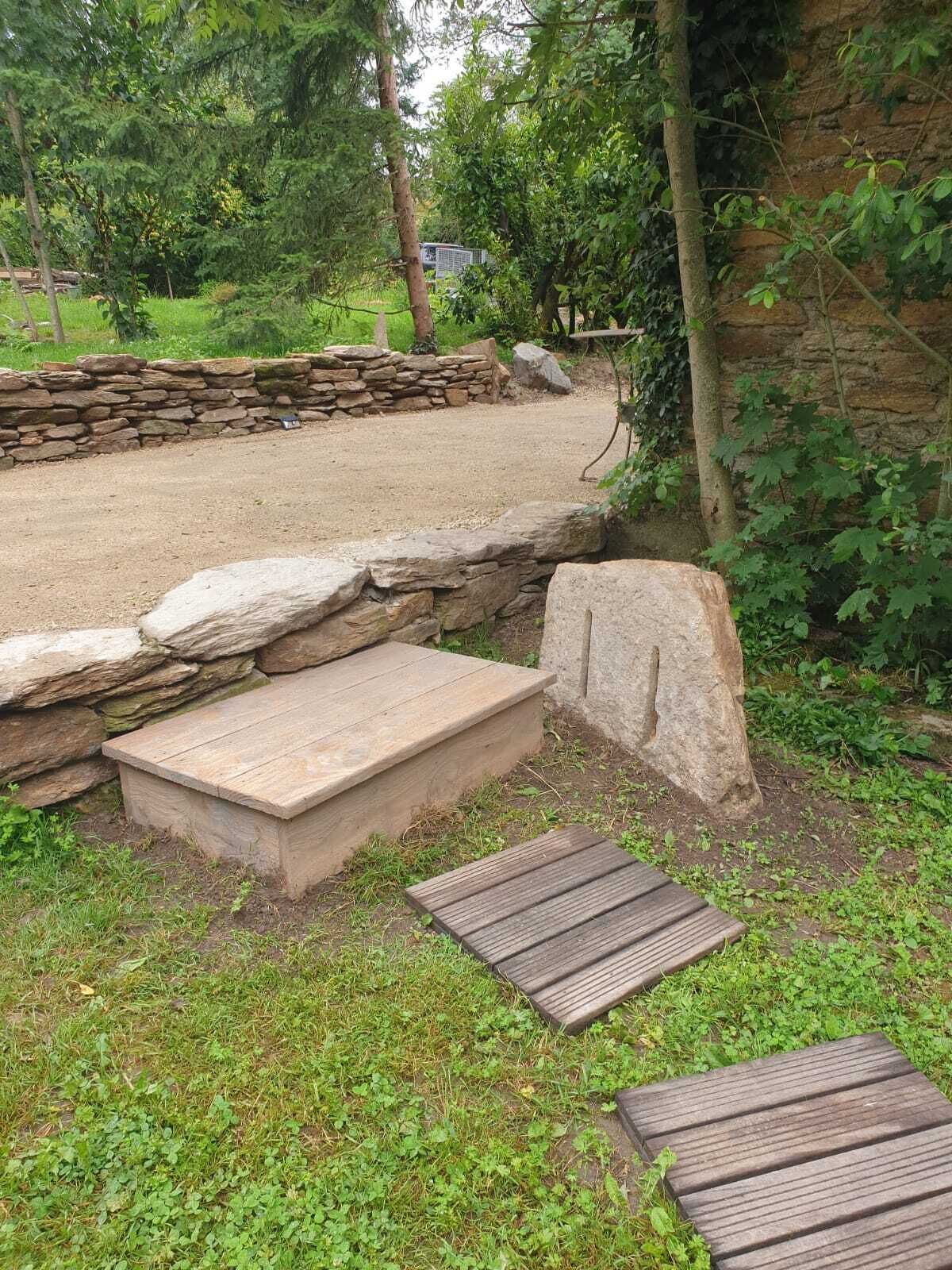 Un chemin de pierres mène à un espace agrémenté d’un banc en bois, entouré de verdure et bordé d’un mur de pierres.