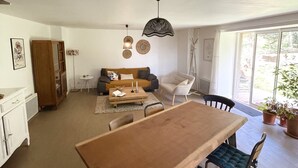 Amplia sala de estar con mesa de comedor de madera, sillas con acolchado azul, sofá y una mesa de centro. Una ventana grande permite la entrada de luz natural.
