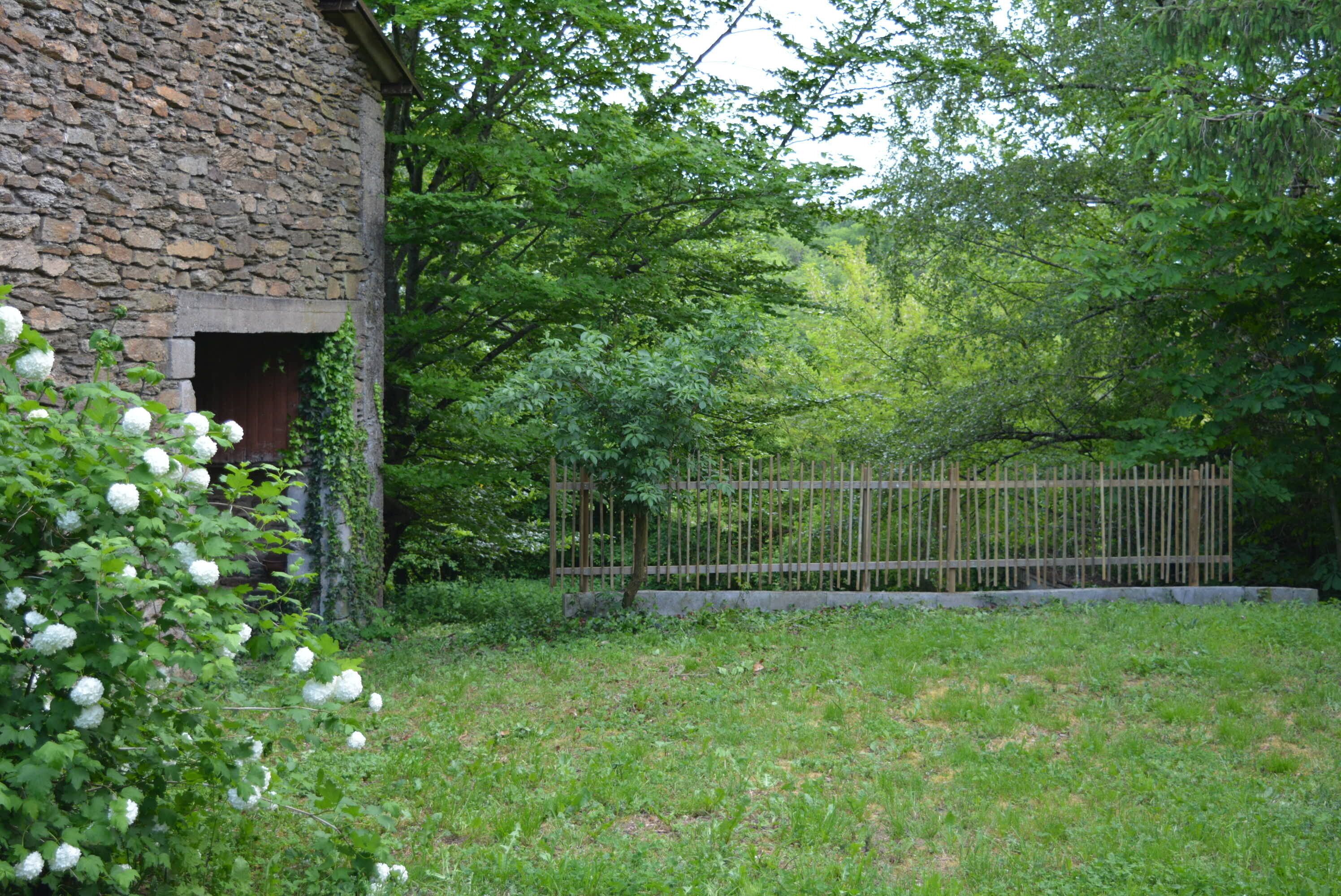 Un bâtiment en pierre avec une porte en bois, entouré d’une végétation luxuriante et d’une clôture en bois.
