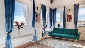 A living room with a green sofa, a small table, and a view of the city through large windows with blue curtains.