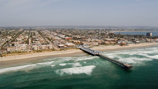 Vista aérea de una ciudad costera con un muelle largo que se adentra en el océano, rodeada de edificios residenciales y una playa.