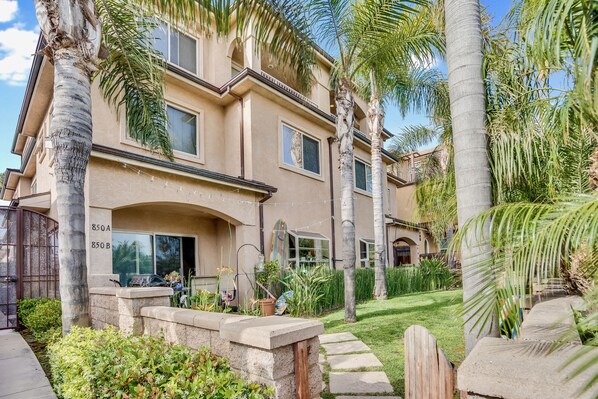 A multi-story building with a beige facade, large windows, and a well-maintained garden with palm trees and shrubs.