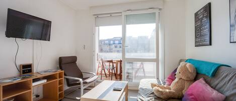 A modern living room with a flat-screen TV, wooden furniture, a sofa with colorful cushions, and a large window offering a view of buildings outside.