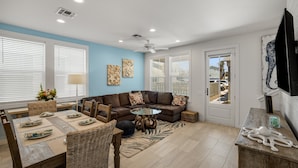 A modern living room with a sectional sofa, a glass coffee table, and a dining area with a wooden table and wicker chairs. - Sea Glass Cottage | Walk to Town! (Port Aransas)