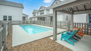 A swimming pool with a wooden deck, surrounded by a wooden fence and a covered seating area with chairs. - Sea Glass Cottage | Walk to Town! (Port Aransas)