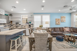 A modern kitchen with a central island, dining area with a glass table, and a living room with a sofa and decorative pillows. - Sea Glass Cottage | Walk to Town! (Port Aransas)