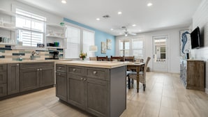 A modern kitchen with a central island, built-in cabinets, and a dining area with a table and chairs. - Sea Glass Cottage | Walk to Town! (Port Aransas)