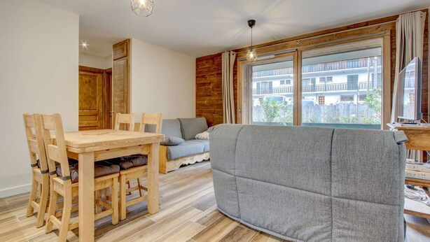 A cozy living area with a wooden dining table, grey sofa, and large windows offering a view of a residential building. - Lapia 3 : New 2 bedrooms with garden by the centre in a tranquil location (MORZINE)