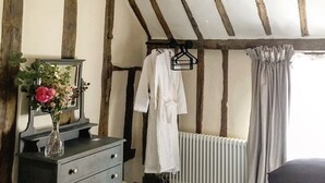 Quarto com vigas de madeira aparente, cômoda com um vaso de flores, roupão pendurado e radiador.