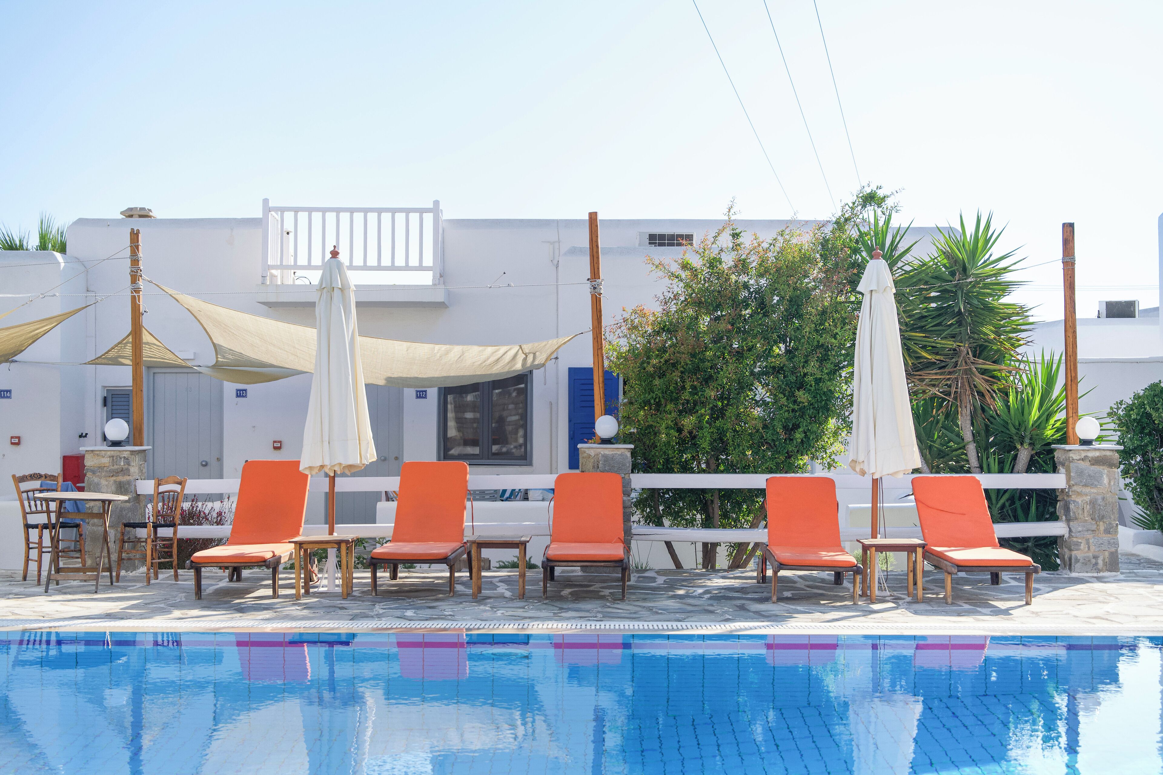 Un espace aménagé au bord de la piscine, avec des chaises longues orange et des parasols blancs, un bâtiment blanc aux volets bleus, et un ciel d’un bleu limpide.
