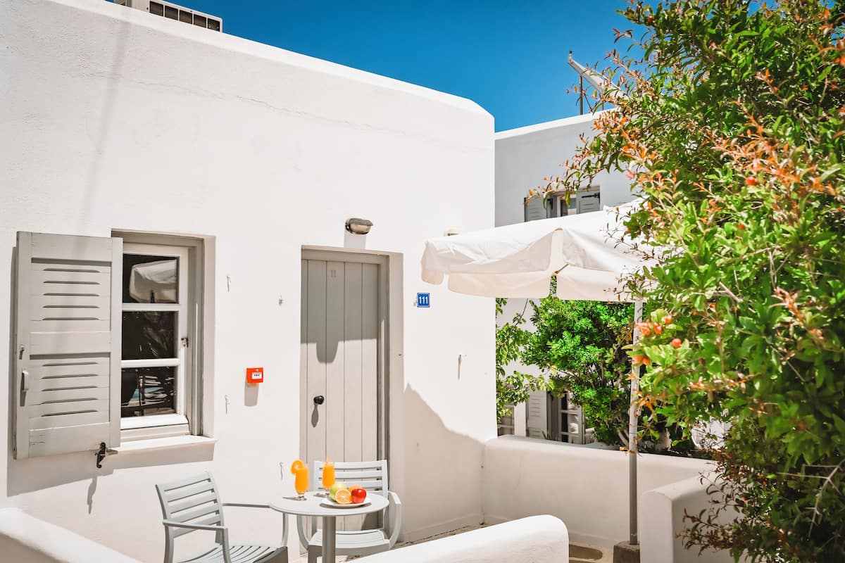 Un edificio blanco con una puerta, una ventana con contraventanas y una mesa con una silla y una cesta de fruta.