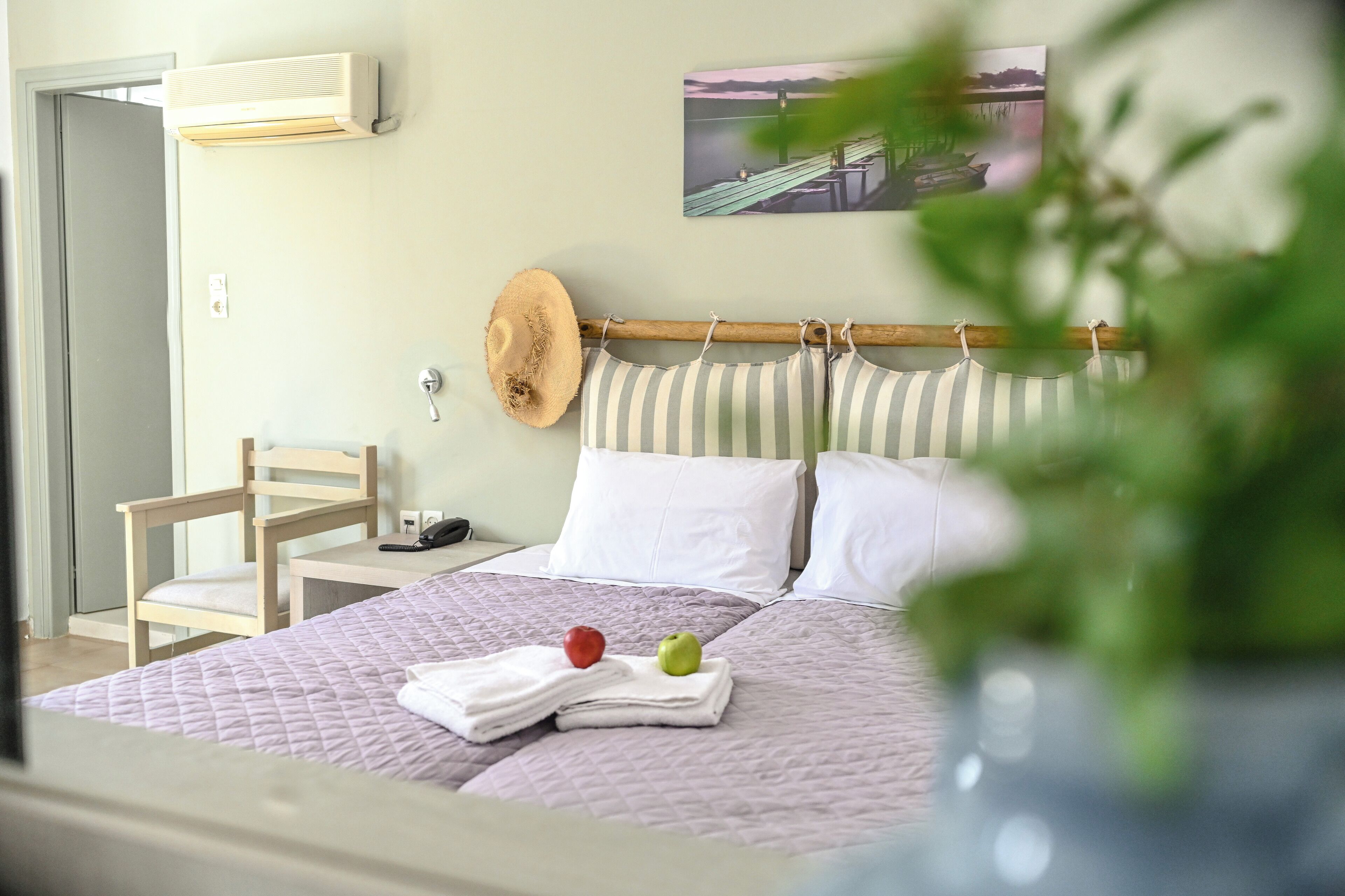 Una habitación de hotel con una cama, una silla, un sombrero, un almohadón y un cuadro en la pared.