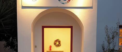 A building entrance with a sign reading "Chambre de Sade." There is a red door, a small shelf with a wreath, and potted plants at the entrance.