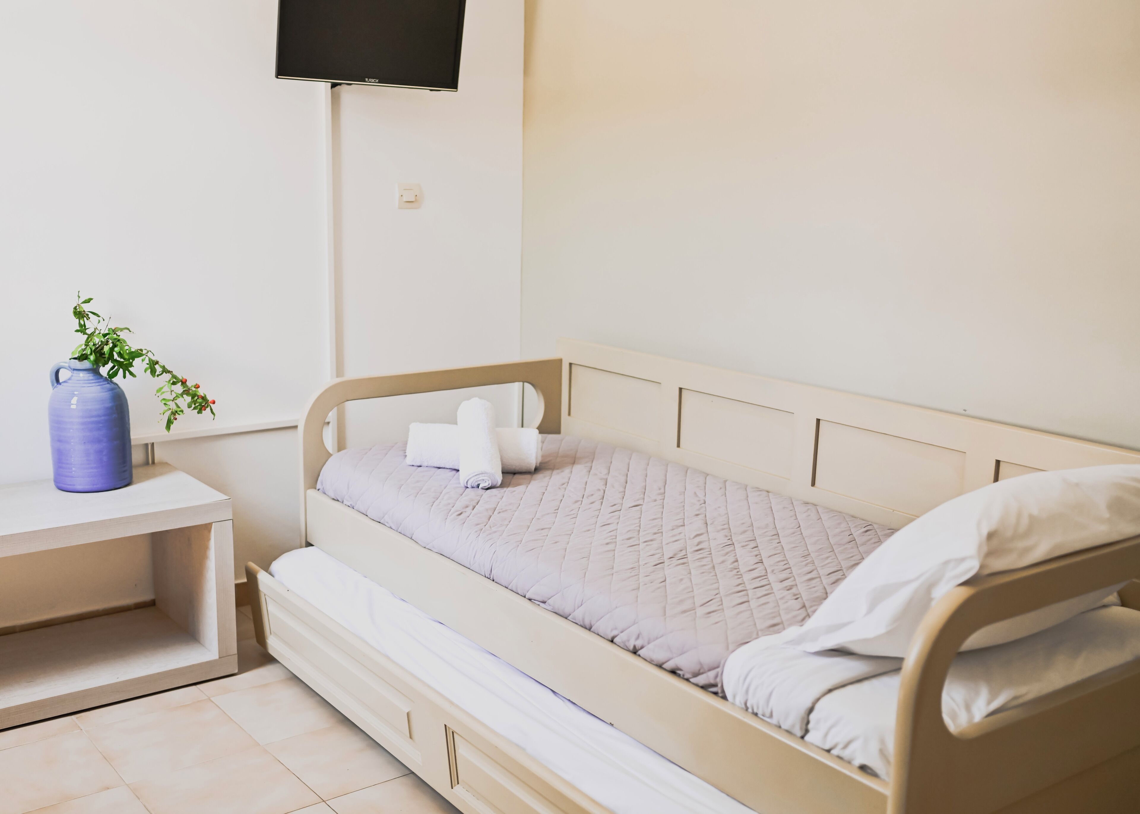 A single bed with a quilted mattress, a bedside table with a vase, and a wall-mounted TV.