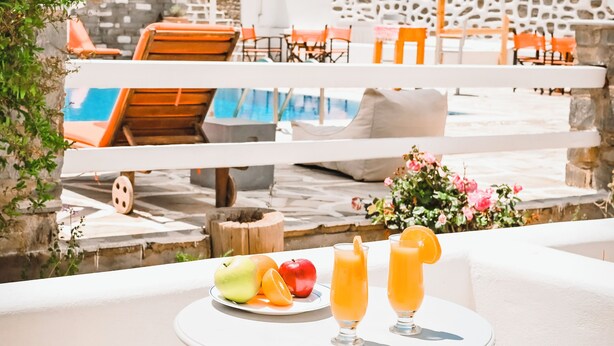 A table with a fruit plate and two glasses of orange juice, outdoor seating with white umbrellas, and a view of a pool and lounge chairs. - Summer Shades (Paros)