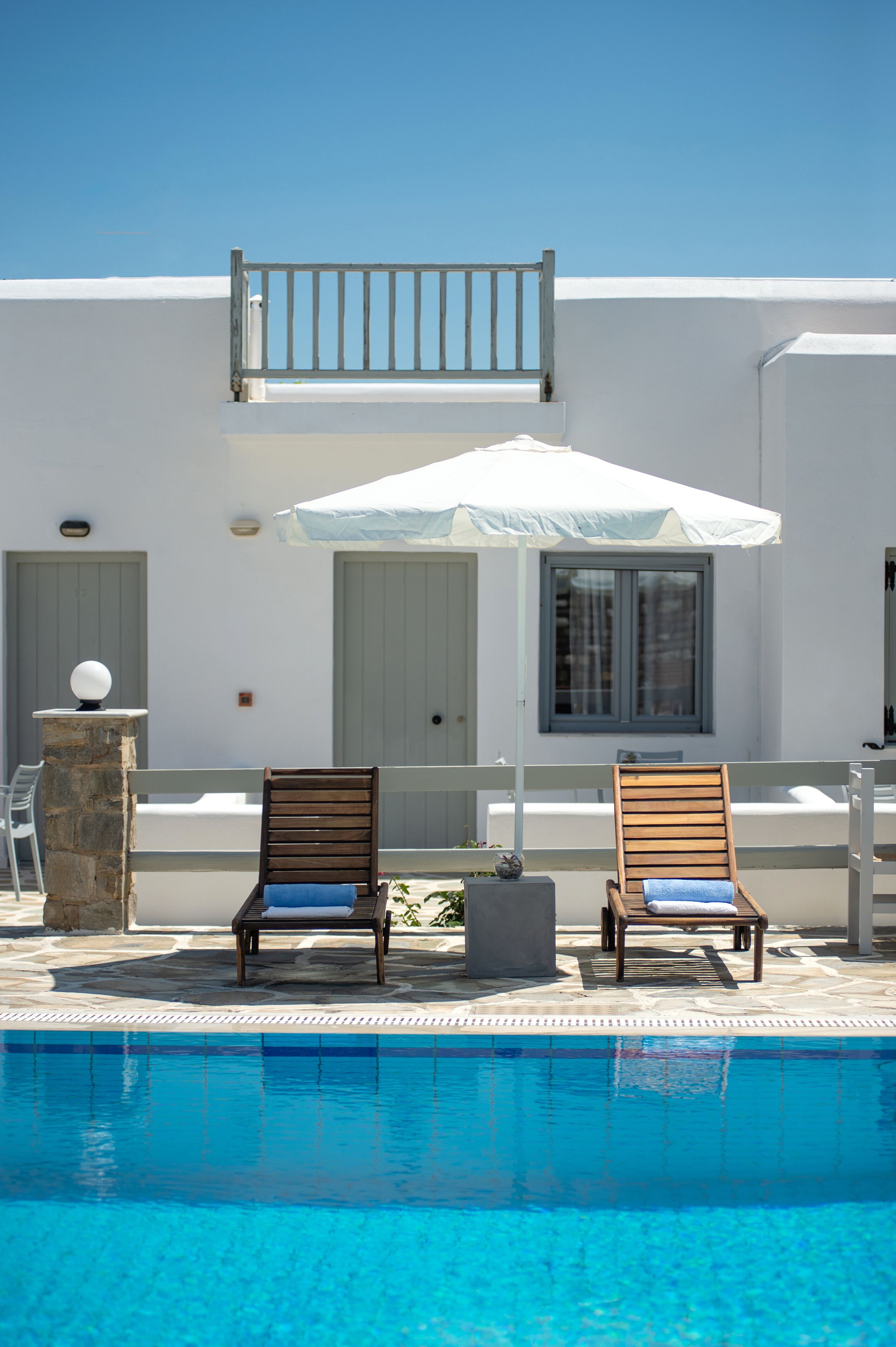 Un espace aménagé au bord de la piscine, comprenant deux chaises longues en bois, un parasol blanc et un pilier en pierre.