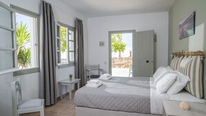 A bedroom with a large bed, a chair, a small table, and a view of the outside. - Summer Shades (Paros)