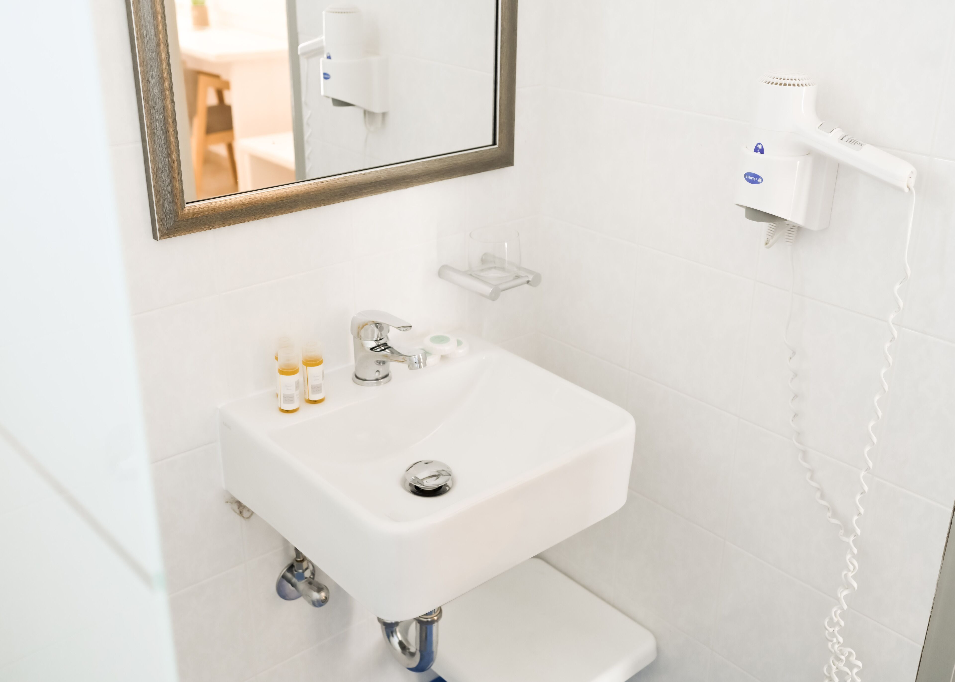 Un baño blanco con lavamanos, dos botellas de loción, un espejo y un secador de pelo en la pared.