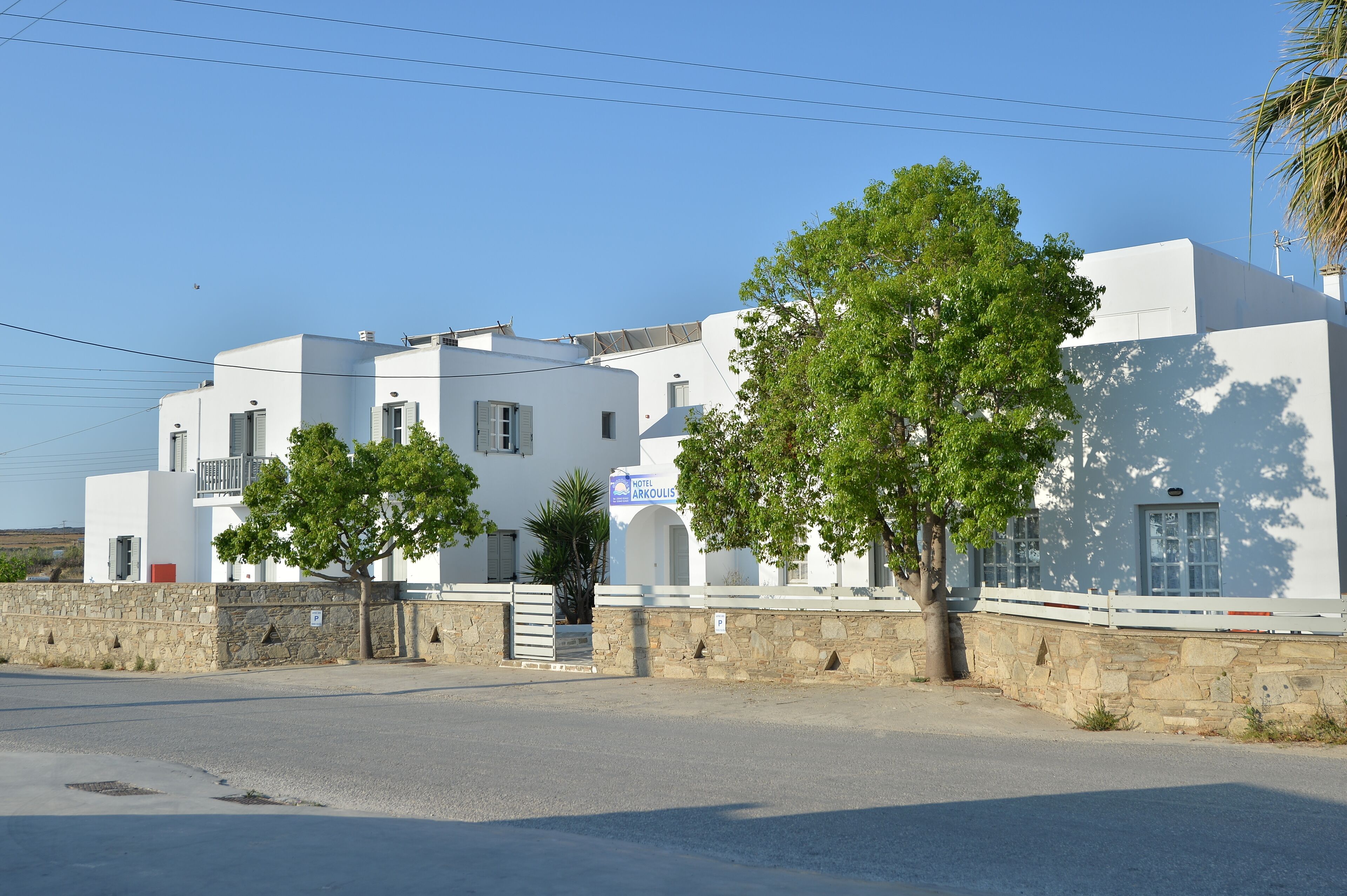 A white building with a sign for Hotel Arkoulis, featuring a stone wall, trees, and a clear sky.