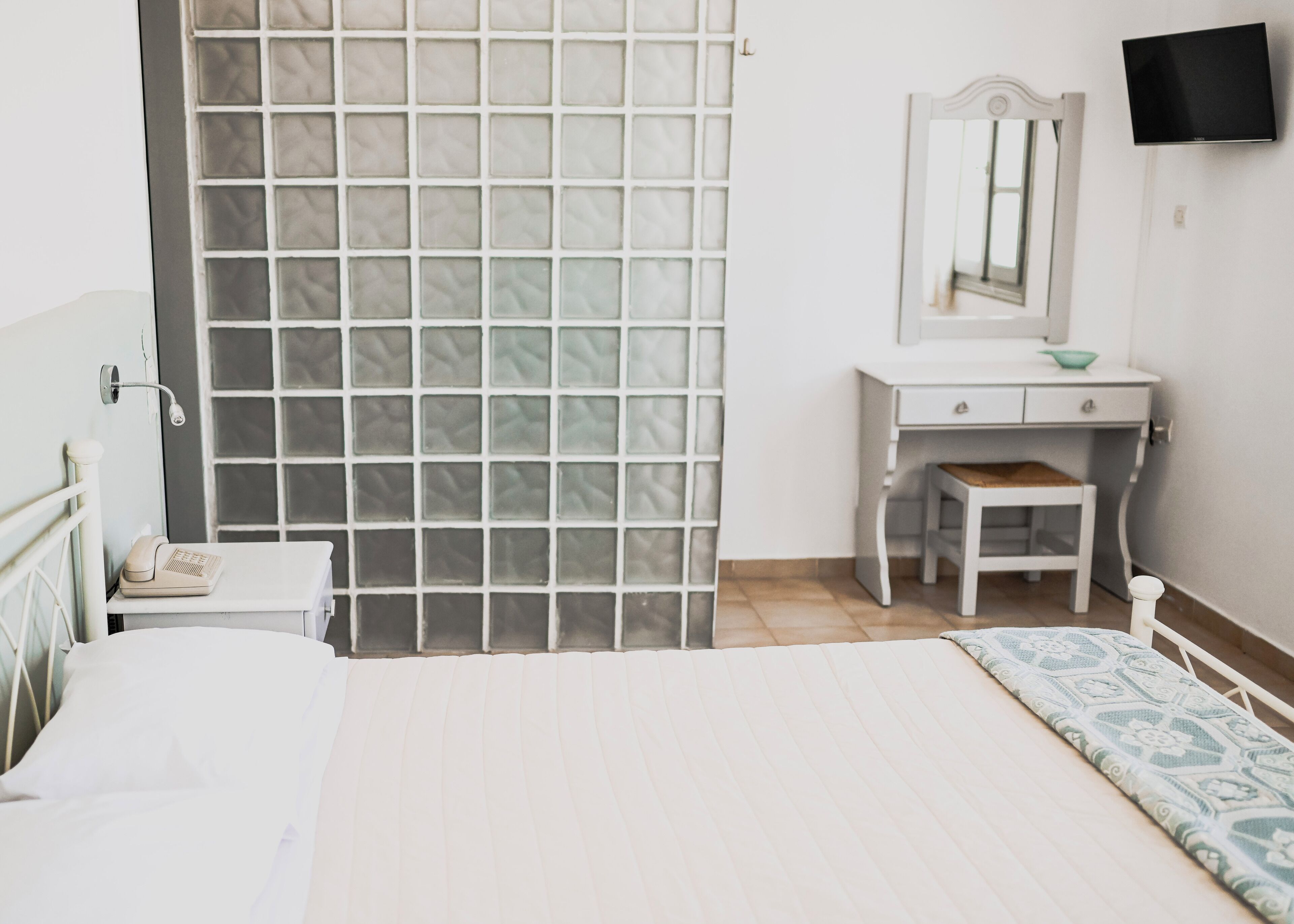 A bedroom with a bed, a bedside table, a small desk with a mirror, and a TV mounted on the wall.