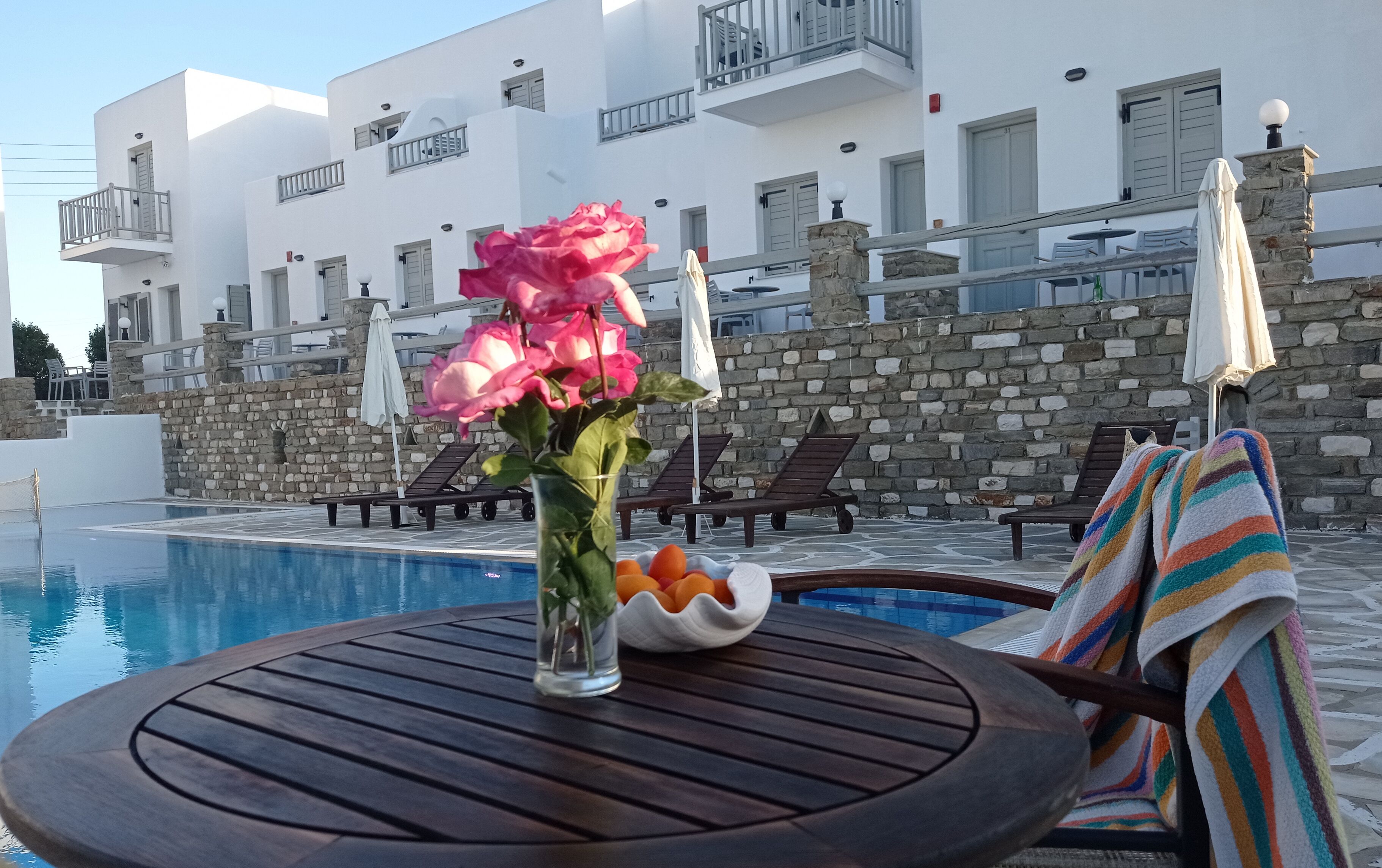 A poolside area with a wooden table, a vase with flowers, a bowl of fruit, and lounge chairs.