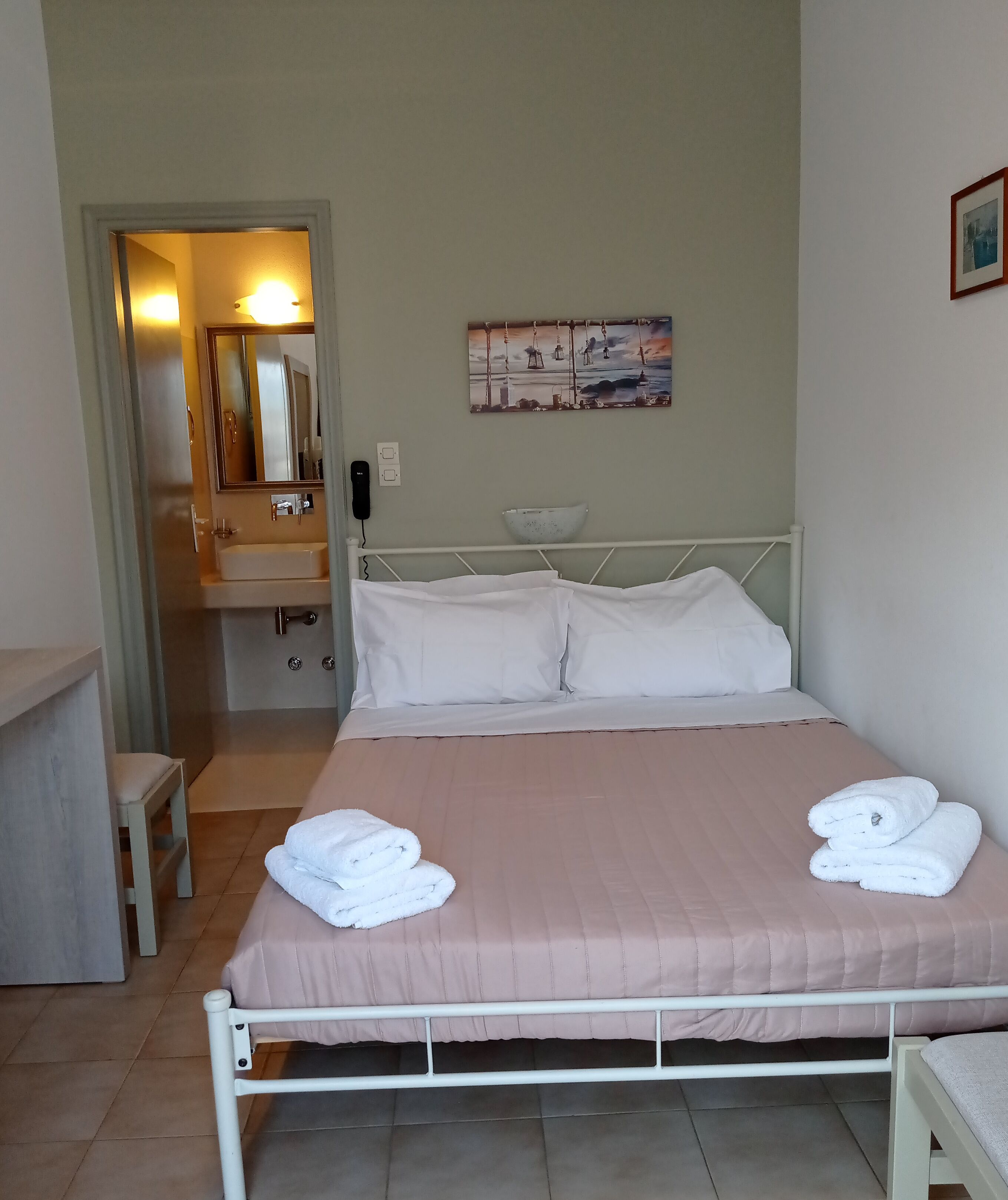 A neatly arranged bedroom with a bed, pillows, a bedside table, a chair, a bathroom visible through an open door, and a painting on the wall.