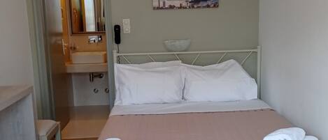 A neatly arranged bedroom with a bed, pillows, a bedside table, a chair, a bathroom visible through an open door, and a painting on the wall.