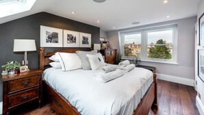 A bedroom with a large bed, a nightstand, a dresser, and a window with a city view. - Luxurious Wandsworth Home Close to Putney Heath by Underthedoormat (London)